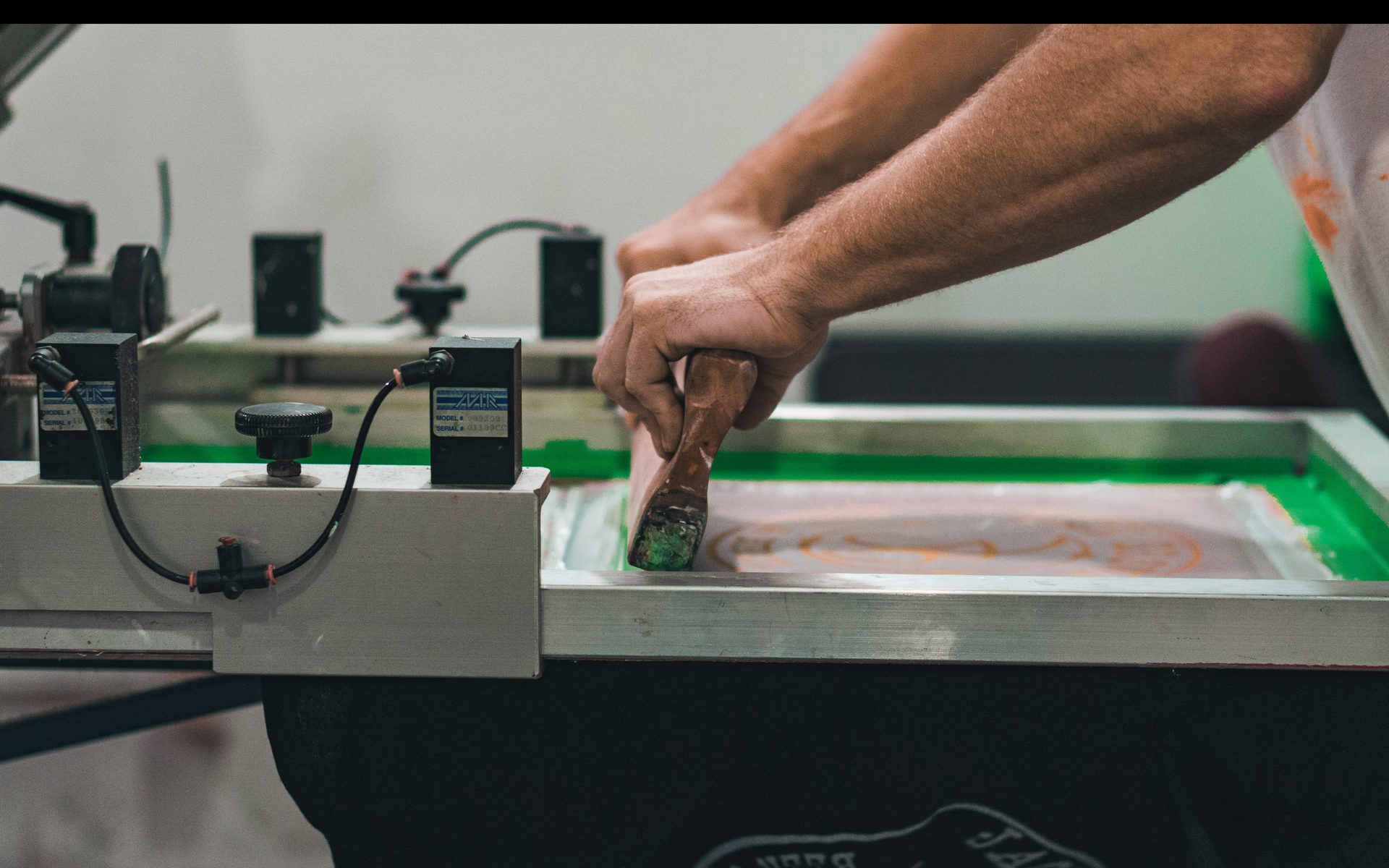 A printer pulling a squeegee by hand on a production-line screen printing press, green ink on mesh